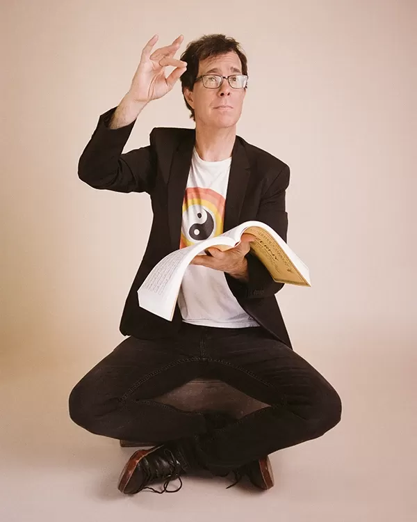 Ben Folds sitting cross-legged, holding a score, and conducting