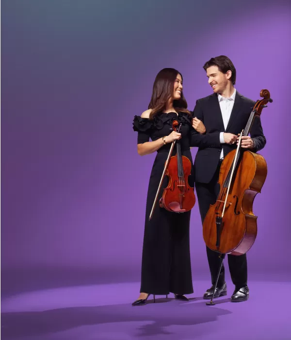 A female violist and a male cellist standind and holding their instruments look lovingly into each other's eyes