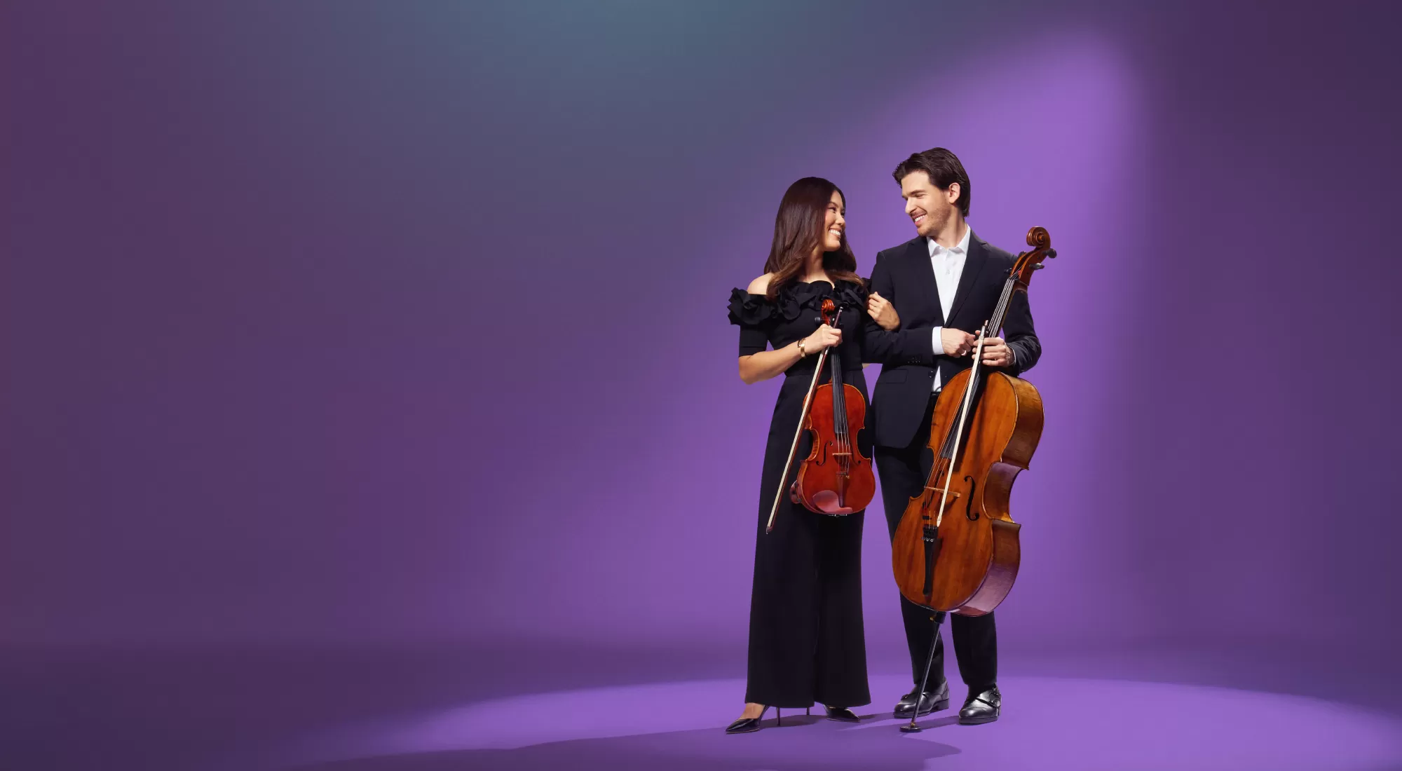 A female violist and a male cellist standind and holding their instruments look lovingly into each other's eyes